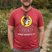 A smiling man stands outdoors on a gravel path, wearing a red heathered t-shirt featuring the Buc-ee's logo—a cartoon beaver in a red hat on a yellow circle—above the text "HUBER HEIGHTS, OH." He has his hands in his pockets, and a pair of sunglasses is tucked into the neckline of the shirt. The background is a lush, slightly blurred Ohio landscape with green trees, autumn foliage, and white wildflowers under an overcast sky.