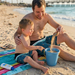A father and young son laughing together on a sunny beach, both wearing matching Buc-ee’s Logo Colorblock swim shorts. The father sits on a colorful beach towel and uses a seashell to pour sand into a solid light blue bucket while his toddler son points and smiles.
