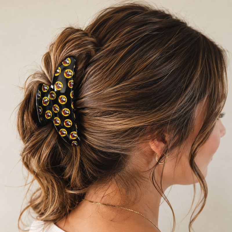 Close-up lifestyle image of a woman wearing a glossy black Buc-ee’s hair claw clip featuring a repeating yellow circular pattern with the Buc-ee’s beaver logo, securing her brown hair in a stylish half-up twist.