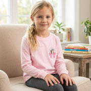 Young girl smiling while sitting on a cozy armchair, wearing the light pink Buc-ee's long-sleeve lounge top with the beaver-in-a-pink-bow logo clearly visible on her chest.