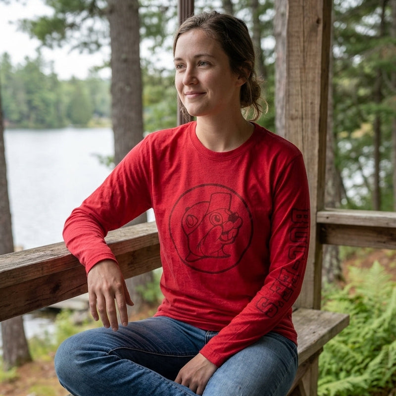 A woman wearing the adult red long-sleeve Buc-ee's beaver logo shirt. She is sitting outdoors on a wooden bench, showcasing the tonal chest logo and the branded left sleeve text. The background is a soft-focus nature scene of a wooded path.
