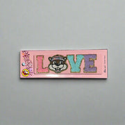 Two Buc-ee’s sticker patches shown on pink packaging: one spells “LOVE” with Buc-ee’s face as the “O,” and the other spells “PEACE” with Buc-ee in the center, both in colorful glitter letters.

