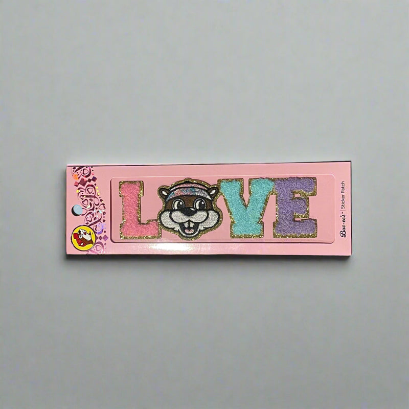 Two Buc-ee’s sticker patches shown on pink packaging: one spells “LOVE” with Buc-ee’s face as the “O,” and the other spells “PEACE” with Buc-ee in the center, both in colorful glitter letters.

