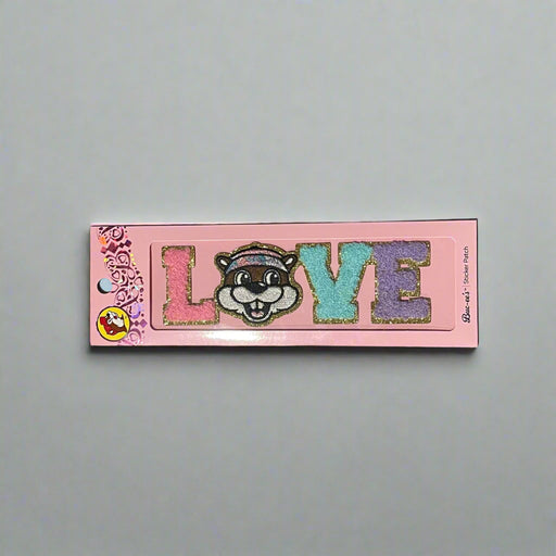 Two Buc-ee’s sticker patches shown on pink packaging: one spells “LOVE” with Buc-ee’s face as the “O,” and the other spells “PEACE” with Buc-ee in the center, both in colorful glitter letters.

