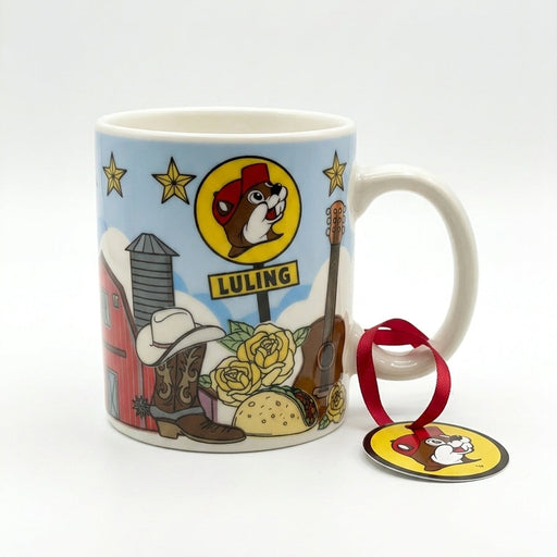 A white ceramic mini mug with a light blue sky backdrop featuring Texas-themed illustrations including a red barn, cowboy boot, taco, guitar, yellow roses, and a Buc-ee's Luling sign. A red hanging ribbon with a circular beaver logo tag is tied to the handle.
