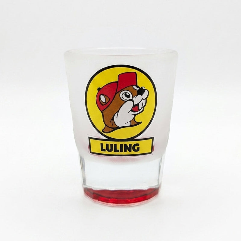A tapered, frosted clear glass shot glass featuring the iconic yellow circular Buc-ee's beaver logo with a yellow banner reading "LULING" underneath. The heavy, solid glass base is tinted in a vibrant, translucent red color.