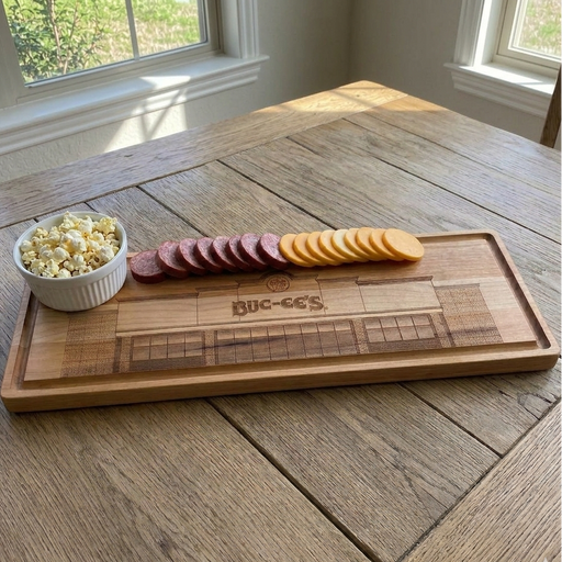 A wide, top-down view of a handcrafted 18-inch by 6-inch Buc-ee's Maple Bar Board sitting on a rustic wooden farmhouse table. The board features a detailed laser engraving of a Buc-ee's storefront and is styled with a small white bowl of popcorn, sliced summer sausage, and cheddar cheese.