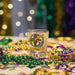 The front view of a colorful Buc-ee's Mardi Gras shot glass featuring Buc-ee the beaver wearing a colorful jester hat with a banner and the year 2026 on it. The shot glass is on a table surrounded by purple, green, and gold beads and confetti, and string lights in the background.