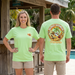 A photograph of two people standing outdoors on a sunny day near a tiki bar and swimming pool, showcasing the front and back of a vibrant lime green t-shirt. On the left, a smiling woman stands facing forward, displaying the front of the shirt with a small tropical logo on the left chest. On the right, a man stands facing away from the camera, clearly displaying the back of the shirt, which features a large circular graphic reading "Brisket Sandwich in Paradise" with a cartoon beaver relaxing in a hammock.