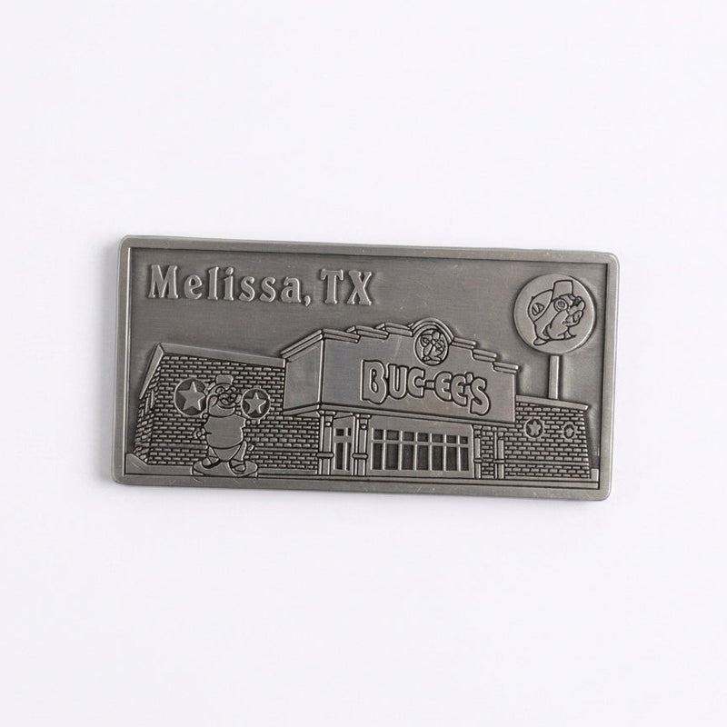 A rectangular pewter metal magnet showing an embossed 3D relief of the Buc-ee's Melissa travel center building, complete with the beaver statue out front, the Buc-ee's sign, a lollipop road sign in the upper right, and the word "Melissa" in the upper left corner.