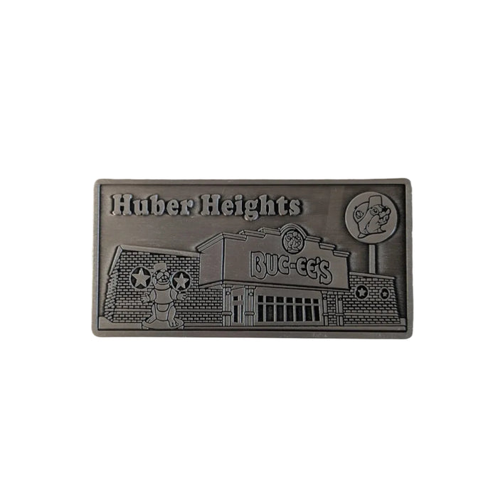 A front-facing photo of a rectangular, embossed metal Buc-ee's magnet for the Huber Heights, Ohio location. The magnet has an antique silver finish and features a 3D relief of the Buc-ee's store building, a beaver mascot statue, and the words "Huber Heights" at the top. It is displayed against a plain white background.