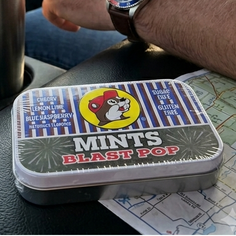 A lifestyle photo taken from inside a truck during a road trip, focusing on a rectangular tin of Buc-ee's Blast Pop Mints resting on a black leather center console next to a paper road map. In the background, a person's arm with a watch rests on the console, a stainless steel travel tumbler sits in the cup holder, and a green Buc-ee's highway sign is visible through the front windshield.