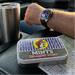 A lifestyle photo taken from inside a truck during a road trip, focusing on a rectangular tin of Buc-ee's Blast Pop Mints resting on a black leather center console next to a paper road map. In the background, a person's arm with a watch rests on the console, a stainless steel travel tumbler sits in the cup holder, and a green Buc-ee's highway sign is visible through the front windshield.