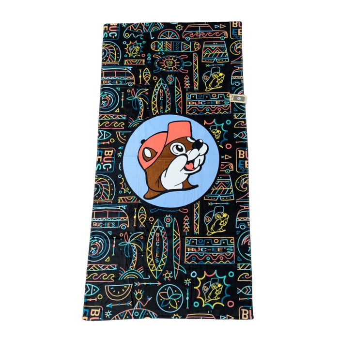 A black beach towel featuring a vibrant "Tiki Neon" pattern with colorful, stylized surfboards, tribal suns, and the Buc-ee's logo in neon blue, orange, and yellow. Displayed on a clean white background.