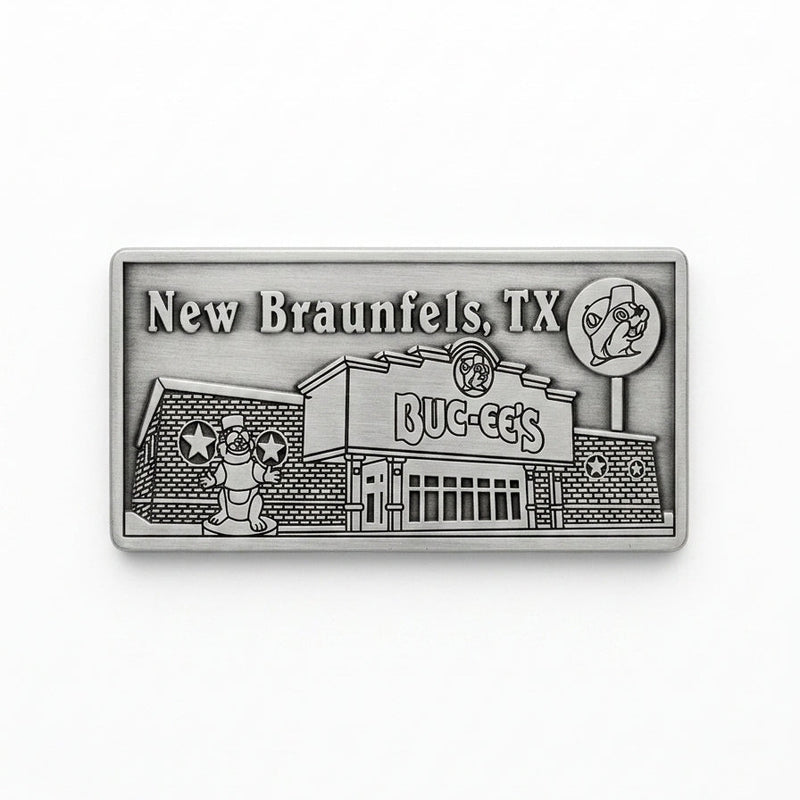 A rectangular pewter metal magnet showing an embossed 3D relief of the Buc-ee's New Braunfels travel center building, complete with the beaver statue out front, the Buc-ee's sign, a lollipop road sign in the upper right, and the word "New Braunfels" in the upper left corner.