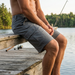 A lifestyle product photo of a man sitting on a rustic wooden dock by a calm lake, showcasing the Buc-ee's Night Owl Fishing Cargo Shorts. The focus is on the right leg of the grey, high-performance shorts, highlighting the large snap-closure cargo pocket and the durable, moisture-wicking fabric. The background features a serene lakeside setting with a forest and a fishing rod resting nearby.