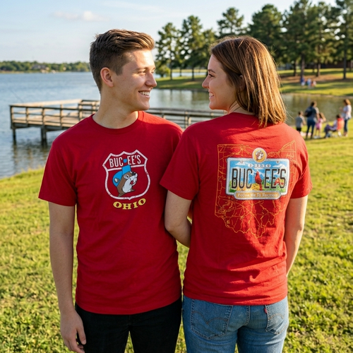 A photo of a smiling young man and woman in red short-sleeve Buc-ee's t-shirts, looking at each other near a lake in the late afternoon. The man, on the left, shows the front of his shirt with the classic white Buc-ee's logo with the beaver and "OHIO" in yellow text. The woman, on the right, shows the back of her shirt with the large license plate logo that reads, "OHIO BUC-EE'S" and has a small graphic of a farm. They stand in front of a blue lake with a wooden pier and pine trees.