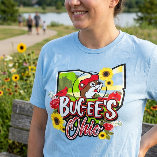 A lifestyle photo of a woman from the chest down, wearing the light blue Buc-ee’s Ohio State Light Shirt. She is smiling and standing in a park with vibrant sunflowers and a walking path in the blurred background. The shirt's central graphic is clearly visible, featuring the Ohio state map, the Buc-ee's beaver mascot, a red barn, and floral accents with "Buc-ee's Ohio" written in bold lettering. 