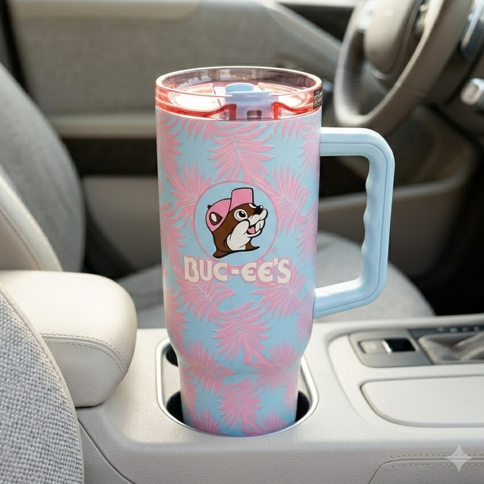 A lifestyle image of a 40-ounce Buc-ee's Pink Palm Fit Forty Pro Tumbler sitting securely in the center console cup holder of a modern vehicle. The tumbler features a light blue background adorned with a continuous pink palm leaf pattern and the iconic circular Buc-ee's beaver mascot logo on the front. It is equipped with a large, light blue ergonomic side handle, a translucent pink lid, and a light blue flip-top closure. The background shows a clean car interior.