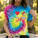 A close-up photograph of a person, from the chest down, wearing a vibrant multi-colored spiral tie-dye t-shirt. The shirt features a blue peace sign, a red heart, and a yellow South Carolina map with a beaver character, above the words "PEACE LOVE S.CAROLINA." The person is sitting on a blanket, holding a simple ceramic mug, outdoors near a tree.
