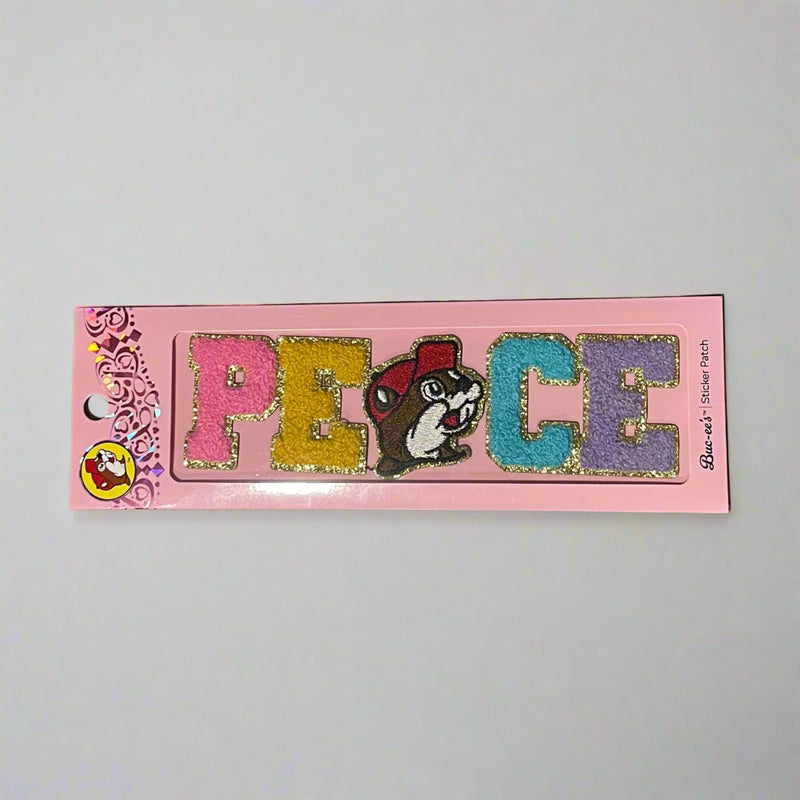 Two Buc-ee’s sticker patches shown on pink packaging: one spells “LOVE” with Buc-ee’s face as the “O,” and the other spells “PEACE” with Buc-ee in the center, both in colorful glitter letters.

