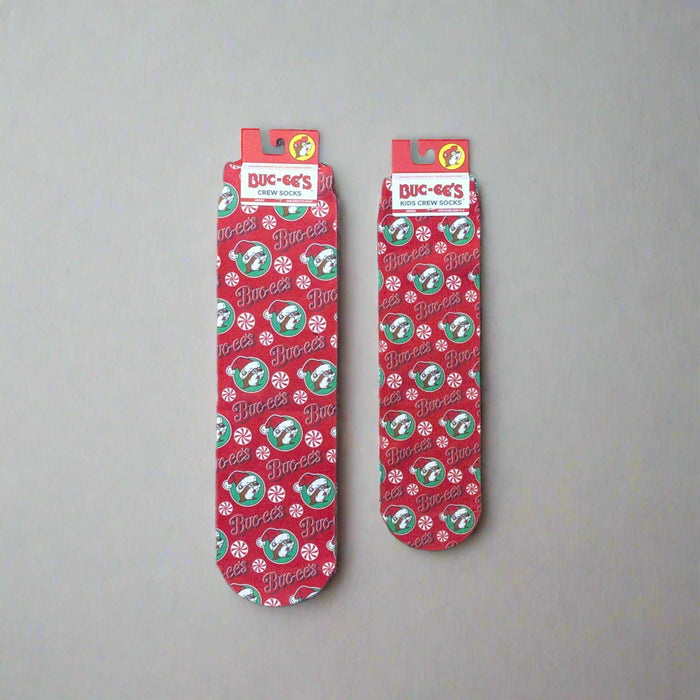 Two pairs of Buc-ee’s Peppermint Christmas Crew Socks, one kids’ size and one adult size, featuring a red background with candy canes, peppermints, and Buc-ee’s logos.