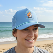 A close-up photograph of a smiling woman wearing the light blue Buc-ee's performance hat. The cap clearly shows the "Buc-ee's Original" beaver patch on the front, the mint green accent rope, and the laser-perforated side panels. The background is a softly blurred beach