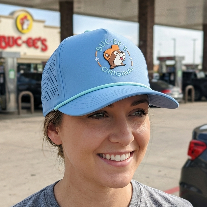 A close-up photograph of a smiling woman wearing the light blue Buc-ee's performance hat. The cap clearly shows the "Buc-ee's Original" beaver patch on the front, the mint green accent rope, and the laser-perforated side panels. The background is a softly blurred Buc-ee's gas station.