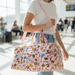 A lifestyle shot of a woman walking through a bright, modern airport terminal carrying the Buc-ee’s Pink Snacks Puffy Duffle Bag. The bag is made of a light pink, quilted "puffy" fabric and features an all-over print of Beaver Nuggets, pretzels, gummy bears, and beverages. She is wearing the bag over her shoulder using a detachable pink strap, alongside the dual top carry handles. The woman is dressed in a simple white t-shirt and blue jeans, with the sunlit terminal and blurred travelers in the background.