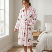 A smiling woman is standing and wearing a Buc-ee's Cherry Plush Robe. A long-sleeved, plush white and pink-striped bathrobe in a brightly lit bedroom. The robe is decorated with a pattern of a Buc-ee beaver wearing a green hat and pink cherries and bows. She is wearing white slippers and is standing next to a window and a bed with a book on it.