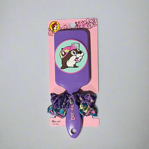 Two Buc-ee’s hair brush and scrunchie sets on display: one with a purple brush and heart scrunchies, the other with a black brush and colorful red-cap Buc-ee's theme scrunchies.

