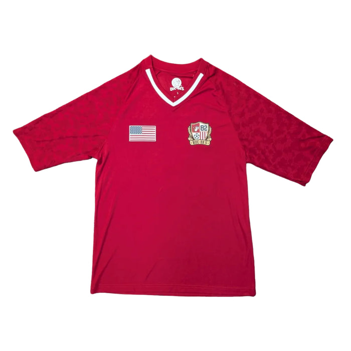 Front view of a Buc-ee’s Red Camo Soccer Jersey featuring tonal red camo sleeves, a white and red ribbed V-neck collar, and a printed American flag on the right chest and a Buc-ee’s '82' soccer crest on the left chest. Displayed on a clean white background.