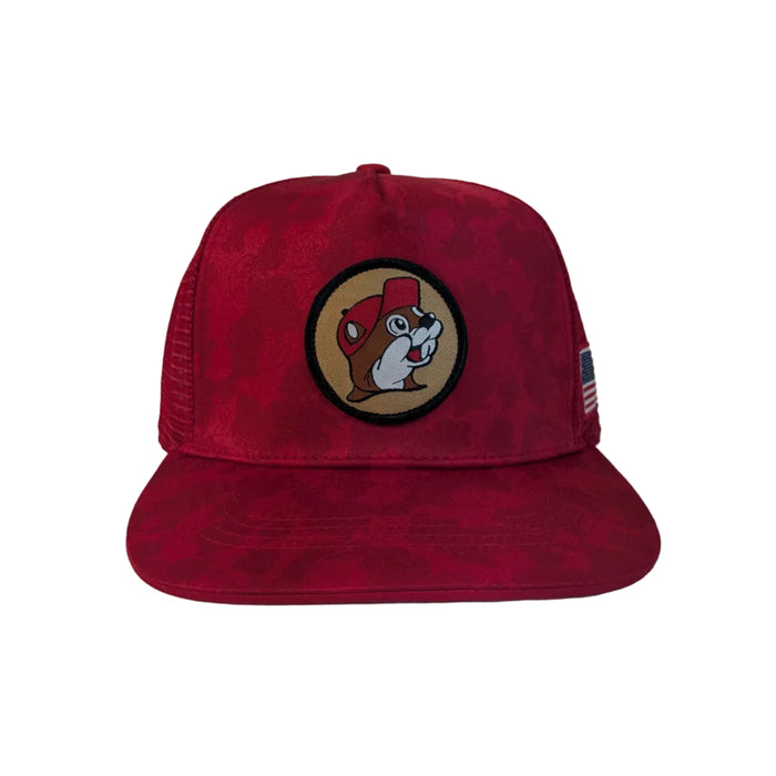 A product shot of a Buc-ee's Red Camo Trucker Hat displayed on a clean white background. The hat features a prominent, circular Buc-ee’s logo patch on the front crown, a tonal red camo pattern on the front panels and brim, and a crisp American flag patch on the side mesh panel.