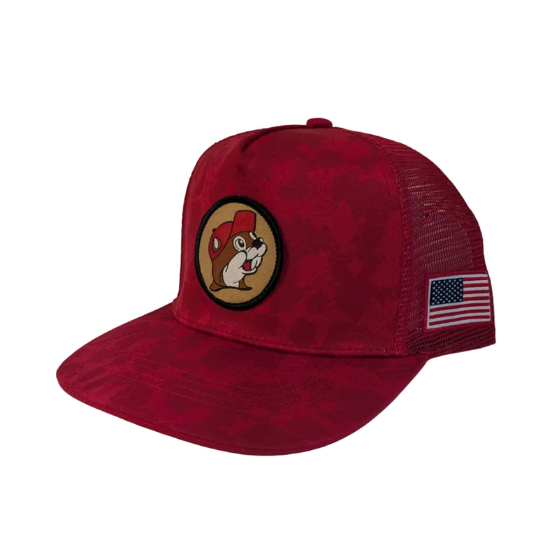Close-up, side profile view of a red camo trucker hat featuring a detailed, rectangular American flag patch embroidered on the side mesh panel, with the red tonal camo pattern visible on the front crown and brim. A circular Buc-ee's beaver logo patch on the crown of the hat. Displayed on a clean white background.