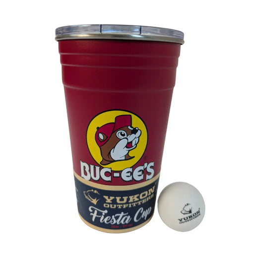 A 20-ounce matte red stainless steel tumbler shaped like a classic party cup with horizontal ridges. It features the Buc-ee's beaver mascot in a yellow circle on the front. A clear lid with a fixed, open drink port is on top, and a white Yukon Outfitters ping pong ball sits on the table next to the cup. The base has a removable navy blue and cream "Fiesta Cup" paper wrap. Displayed on a clean white background.
