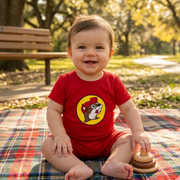 A happy baby with a cheerful expression sits on a multi-colored plaid picnic blanket in a sun-drenched park. The baby is wearing a bright red, short-sleeve Buc-ee's romper featuring the iconic yellow beaver logo on the chest. The romper has a smooth crew neckline and short-style legs. In the soft-focus background, there is a wooden park bench, green grass, and leafy trees under a bright sky. A wooden stacking toy sits on the blanket next to the baby.