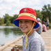 A lifestyle photo of a smiling youth boy sitting on a wooden fishing pier by a calm lake. He is wearing the Buc-ee’s Red Reversible Kids' Bucket Hat, which features a vibrant red crown, a yellow contrast band with "BUC-EE'S" text, and the circular beaver logo on the front. The underside of the brim is a contrasting royal blue. The boy is holding a fishing rod, with a tackle bag and colorful bobbers visible on the pier beside him.