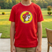 Young boy wearing a red t-shirt with a logo, standing on a patio with a grassy area and trees in the background.