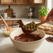 A hand holding a Buc-ee's Spatula featuring a wooden-handled spatula with a black silicone head covered in a yellow beaver logo pattern, lifting melted chocolate batter from a large white ceramic bowl. In the background of the bright kitchen, a second red spatula with a yellow logo-print head sits inside another mixing bowl.