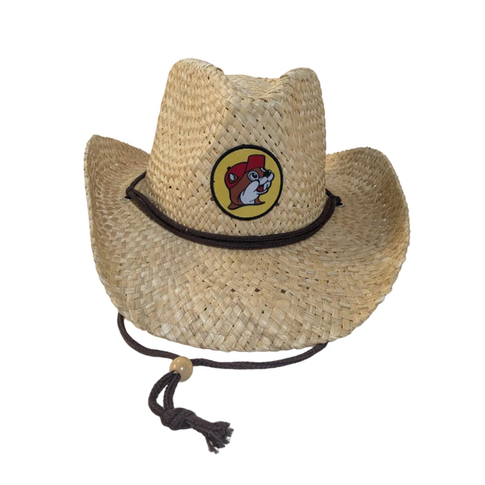 A Buc-ee's River Straw Western Hat. A natural, light-colored woven straw Western-style hat sitting on a white surface. The hat features a prominent circular Buc-ee's beaver logo patch on the front crown, a dark brown decorative hat band, and an attached dark brown chin cord with a sliding wooden bead.