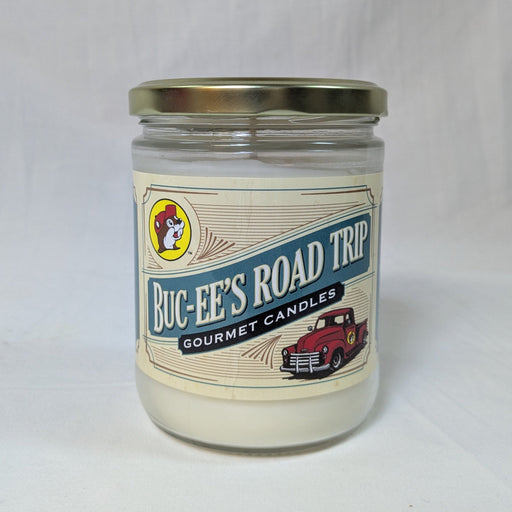 A front-facing product shot of a Buc-ee’s Road Trip Gourmet Candle in a clear glass jar with a gold metallic lid. The vintage-style cream label features the iconic Buc-ee's beaver mascot in a yellow circle on the top left. Large blue block letters read "BUC-EE’S ROAD TRIP," underlined by a black banner that says "GOURMET CANDLES" in white text. An illustration of a classic red pickup truck is featured on the bottom right of the label. The candle is set against a clean, neutral white fabric background.