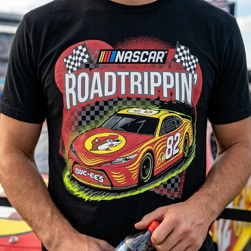 A close-up lifestyle shot of a person wearing a black Buc-ee’s NASCAR 'Road Trippin’' graphic t-shirt. The shirt features a large, detailed illustration of a red and yellow #82 stock car with a Buc-ee’s mascot logo on the hood. Above the car, the word 'NASCAR' is printed in its signature block font next to the colorful stripes, with 'ROAD TRIPPIN’' in bold white letters below it. The background shows a blurred outdoor race track setting.