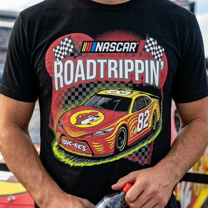 A close-up lifestyle shot of a person wearing a black Buc-ee’s NASCAR 'Road Trippin’' graphic t-shirt. The shirt features a large, detailed illustration of a red and yellow #82 stock car with a Buc-ee’s mascot logo on the hood. Above the car, the word 'NASCAR' is printed in its signature block font next to the colorful stripes, with 'ROAD TRIPPIN’' in bold white letters below it. The background shows a blurred outdoor race track setting.