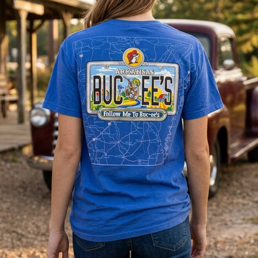 A woman viewed from behind wearing a blue graphic t-shirt with a 'Buck-EE's' license plate design on the back, standing outdoors in front of a rustic wooden building and a vintage truck.