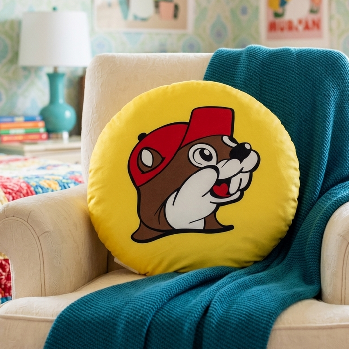 A round, yellow Buc-ee’s logo throw pillow featuring the iconic cartoon beaver wearing a red baseball cap. The pillow sits upright in a cream-colored upholstered armchair. A solid teal, knit blanket is draped over the back and across the right arm of the chair. The background shows a colorful bedroom with patterned wallpaper, framed art prints, a teal table lamp, and a quilted bed.