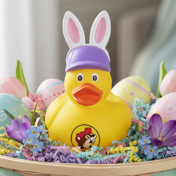A yellow Buc-ee’s rubber ducky with white and pink bunny ears, light purple cap, orange beak and Buc-ee's logo on the front, nestled inside a pastel Easter basket filled with colorful shredded paper, decorated eggs, and spring flowers.