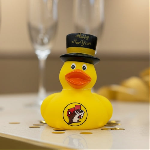 Buc-ee’s yellow rubber ducky wearing a black Happy New Year top hat with Buc-ee’s logo on the front, displayed on a counter with gold confetti around it and champagne flutes blurred in the background.