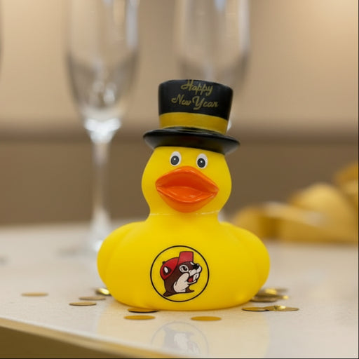 Buc-ee’s yellow rubber ducky wearing a black Happy New Year top hat with Buc-ee’s logo on the front, displayed on a counter with gold confetti around it and champagne flutes blurred in the background.