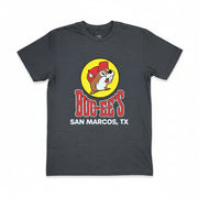 A dark heather gray t-shirt laid flat on a textured gray carpet, featuring a graphic of a beaver in a red cap inside a yellow circle above the red text 'BUC-EE'S' and white text 'SAN MARCOS, TX'.