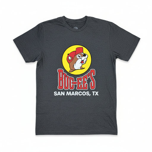 A dark heather gray t-shirt laid flat on a textured gray carpet, featuring a graphic of a beaver in a red cap inside a yellow circle above the red text 'BUC-EE'S' and white text 'SAN MARCOS, TX'.